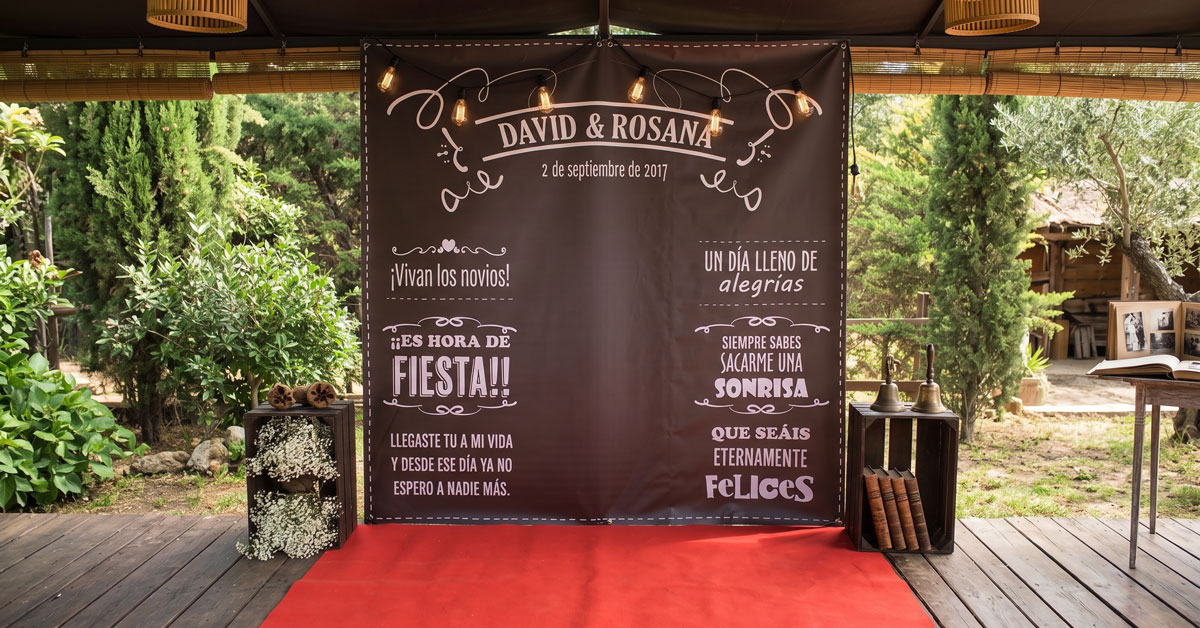 Photocall de boda con los nombres de los novios y la fecha, montado sobre alfombra roja en el porche de una finca, entre cipreses y olivos